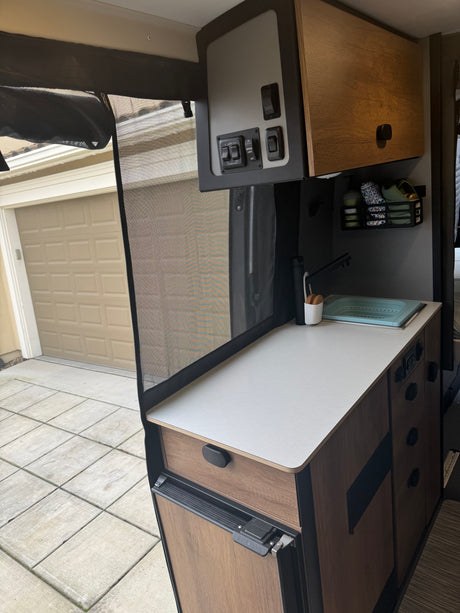 RV rental kitchen setup with white countertop, sink, storage, Nova Kool fridge, and control panel, viewed through a mesh screen.