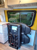 RV rental interior with two black passenger seats with visible folding mechanism next to a window overlooking greenery. Wood details.