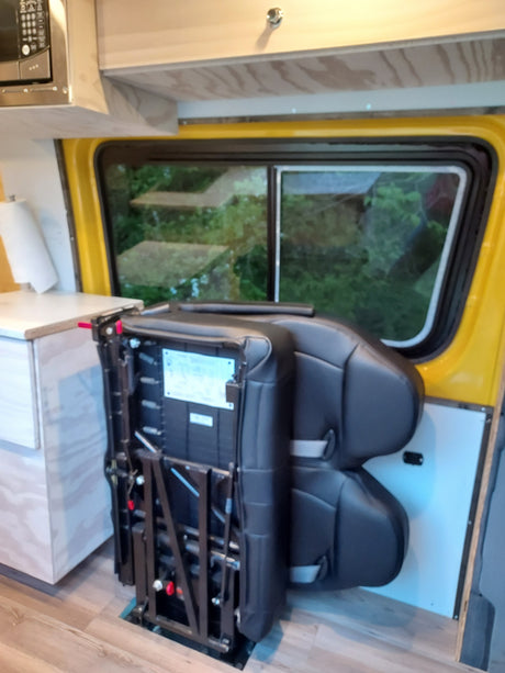 RV rental interior with two black passenger seats with visible folding mechanism next to a window overlooking greenery. Wood details.