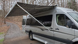 Experience an RV rental: a silver camper van with its striped awning fully extended, parked on a path scattered with pinecones.