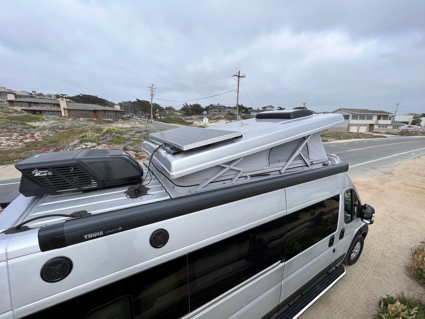 Overhead view of a silver camper van, possibly an rv rental, with a pop-top, solar panel, and Dometic AC unit, parked by a road.