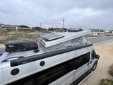 Overhead view of a silver camper van, possibly an rv rental, with a pop-top, solar panel, and Dometic AC unit, parked by a road.