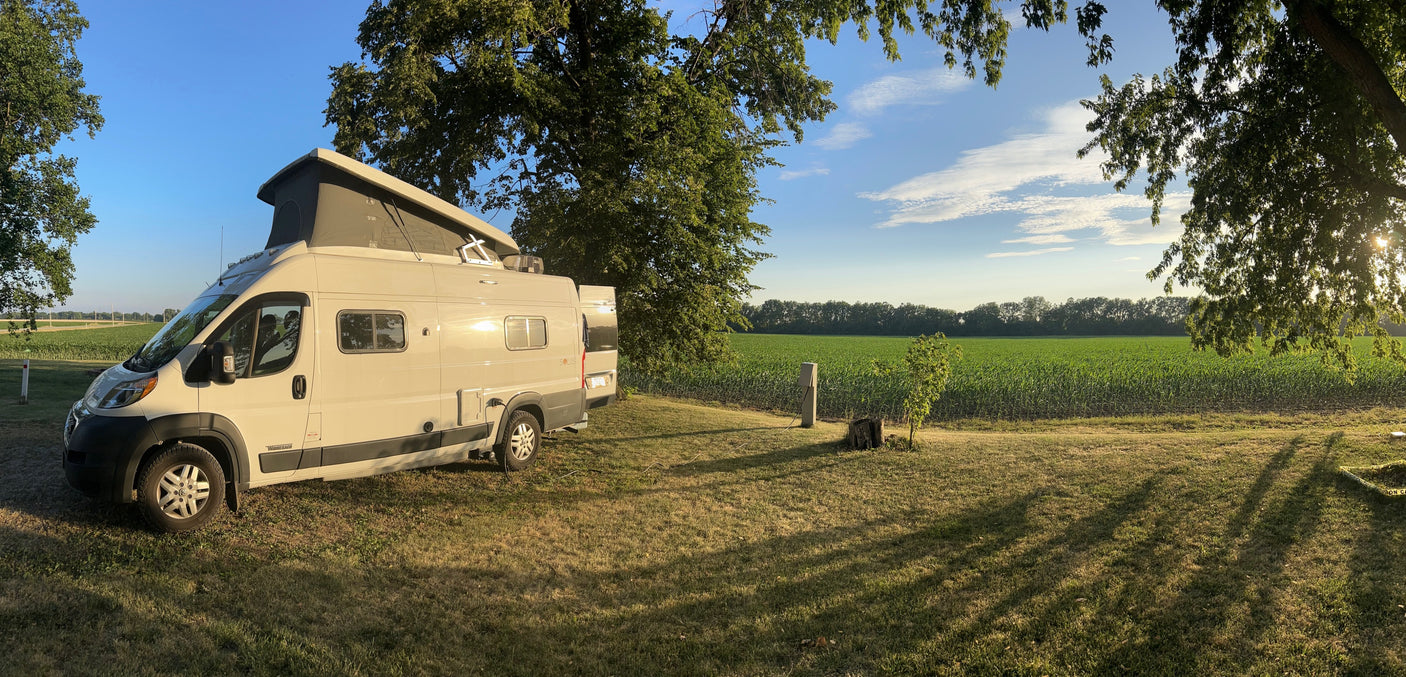 Peaceful RV rental: Beige pop-top camper van on grass with long shadows, facing a green crop field under blue sky, surrounded by trees.