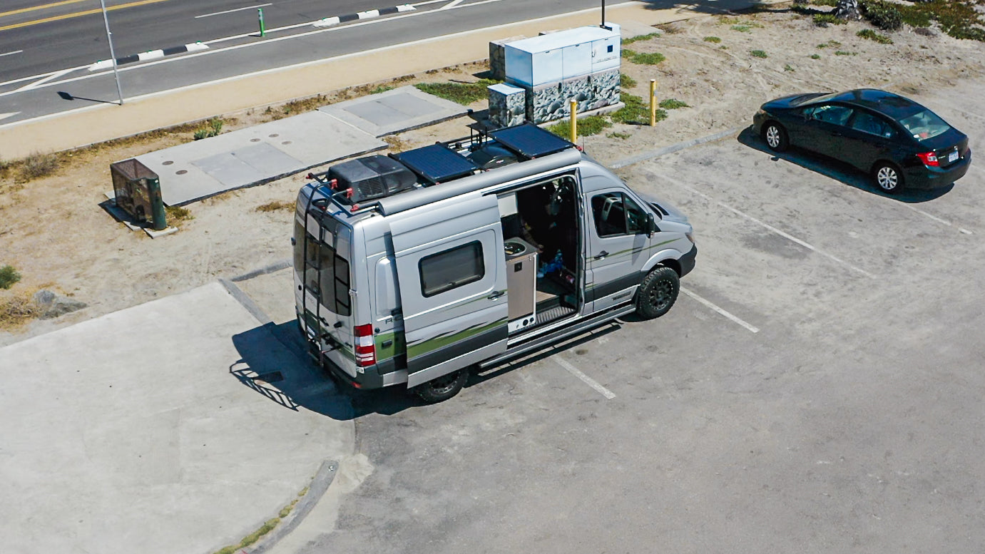 A grey RV rental camper van with rooftop solar panels and open side door, parked in a sunny lot near a road. Ideal for travel.