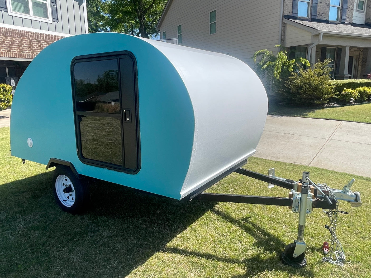 A compact teal & white teardrop camper, perfect for an rv rental, parked on a bright lawn by houses.