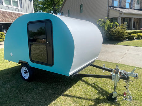 A compact teal & white teardrop camper, perfect for an rv rental, parked on a bright lawn by houses.