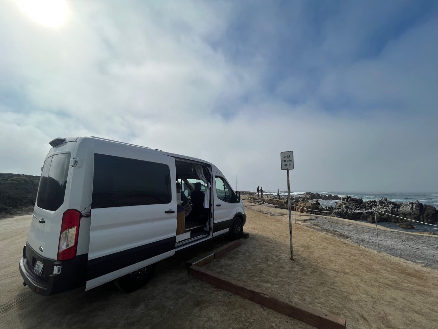 White rv rental van parked on sandy coast by rocky ocean. Open door reveals interior. Angle Parking sign visible for travelers.