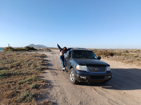 Woman enjoying a desert adventure, posing joyfully by a Ford Expedition SUV on a dirt road. Ideal scenery for an RV rental trip.