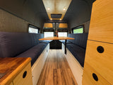 RV rental interior: A bright, modern dinette with blue cushioned benches, a wooden table, and wood floors in a comfy camper van.