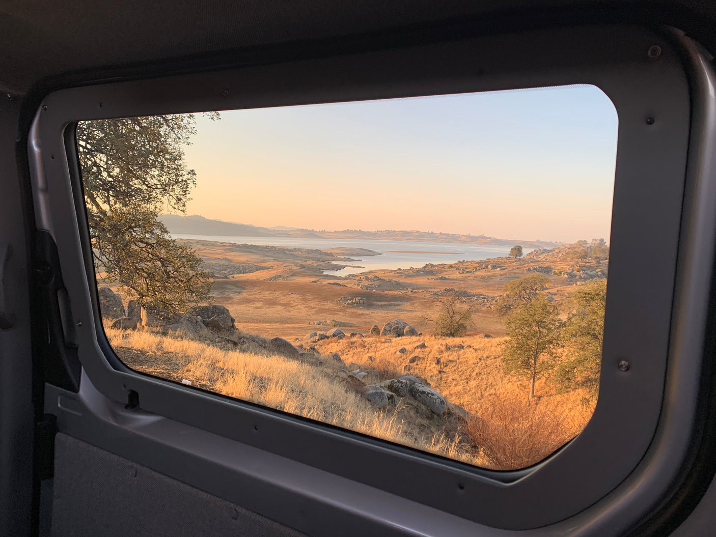 Scenic golden hour view from an RV rental window, showing a tranquil lake, rocky hills, and dry landscape.