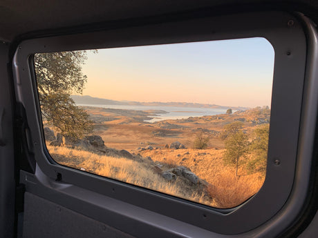 Scenic golden hour view from an RV rental window, showing a tranquil lake, rocky hills, and dry landscape.