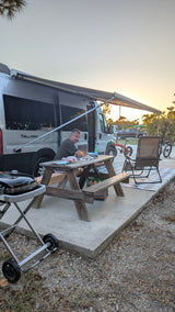 Man relaxing at his RV rental campsite with a picnic table, grill, and camper van under an awning at sunset.