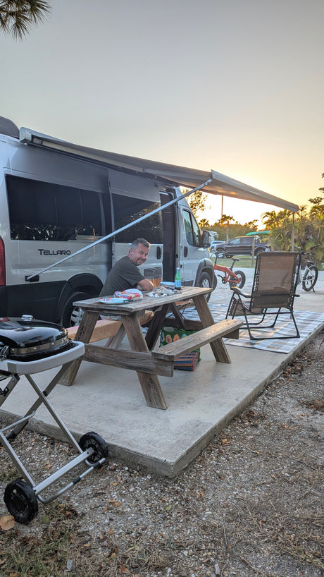 Man relaxing at his RV rental campsite with a picnic table, grill, and camper van under an awning at sunset.