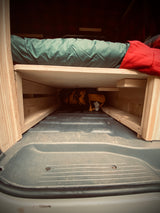 RV rental: Inside a camper van, a custom wooden bed with green sleeping bag & under-bed storage.