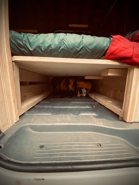 RV rental: Inside a camper van, a custom wooden bed with green sleeping bag & under-bed storage.