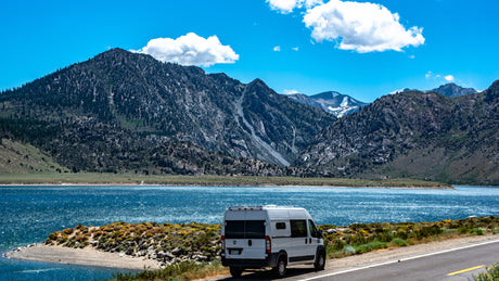 A white RV rental parked on a scenic road beside a shimmering blue lake, with towering mountains and a clear sky.
