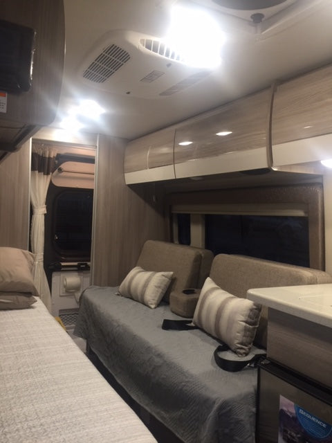 Bright RV rental interior showing a bed, lounge seating, overhead cabinets, and AC unit. A compact modern camper living space.
