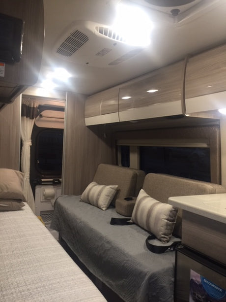 Bright RV rental interior showing a bed, lounge seating, overhead cabinets, and AC unit. A compact modern camper living space.