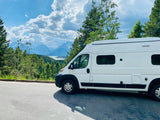 A white rv rental van rests at a scenic overlook, showcasing majestic Grand Teton mountains, a lake, and lush forests under a cloudy sky.