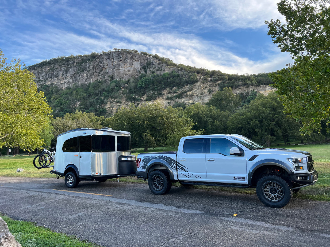 RV rental: a white Ford Raptor truck towing an Airstream trailer with bikes, parked roadside before tall cliffs.