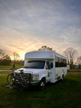 White RV rental with front-mounted bikes, parked on green grass at sunset. Ideal for outdoor adventures.