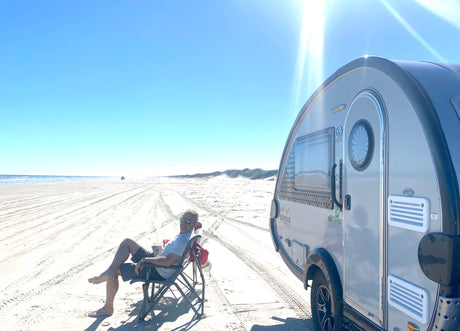 Beach RV rental adventure: A man unwinds in a chair beside his teardrop camper on a vast, sunny sandy beach.