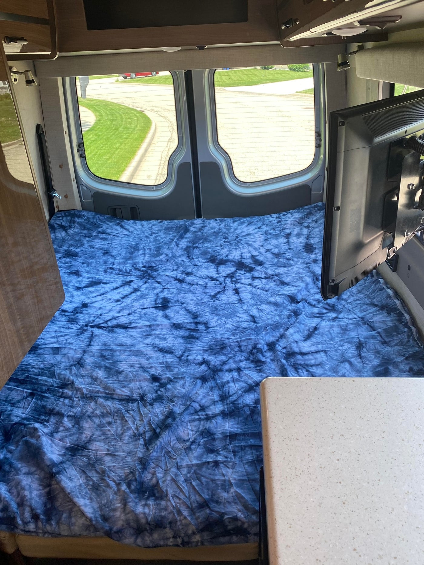Inside an RV rental: a blue tie-dye bed fills the back, with a TV, kitchen counter, and rear windows showing a road. — photo 5