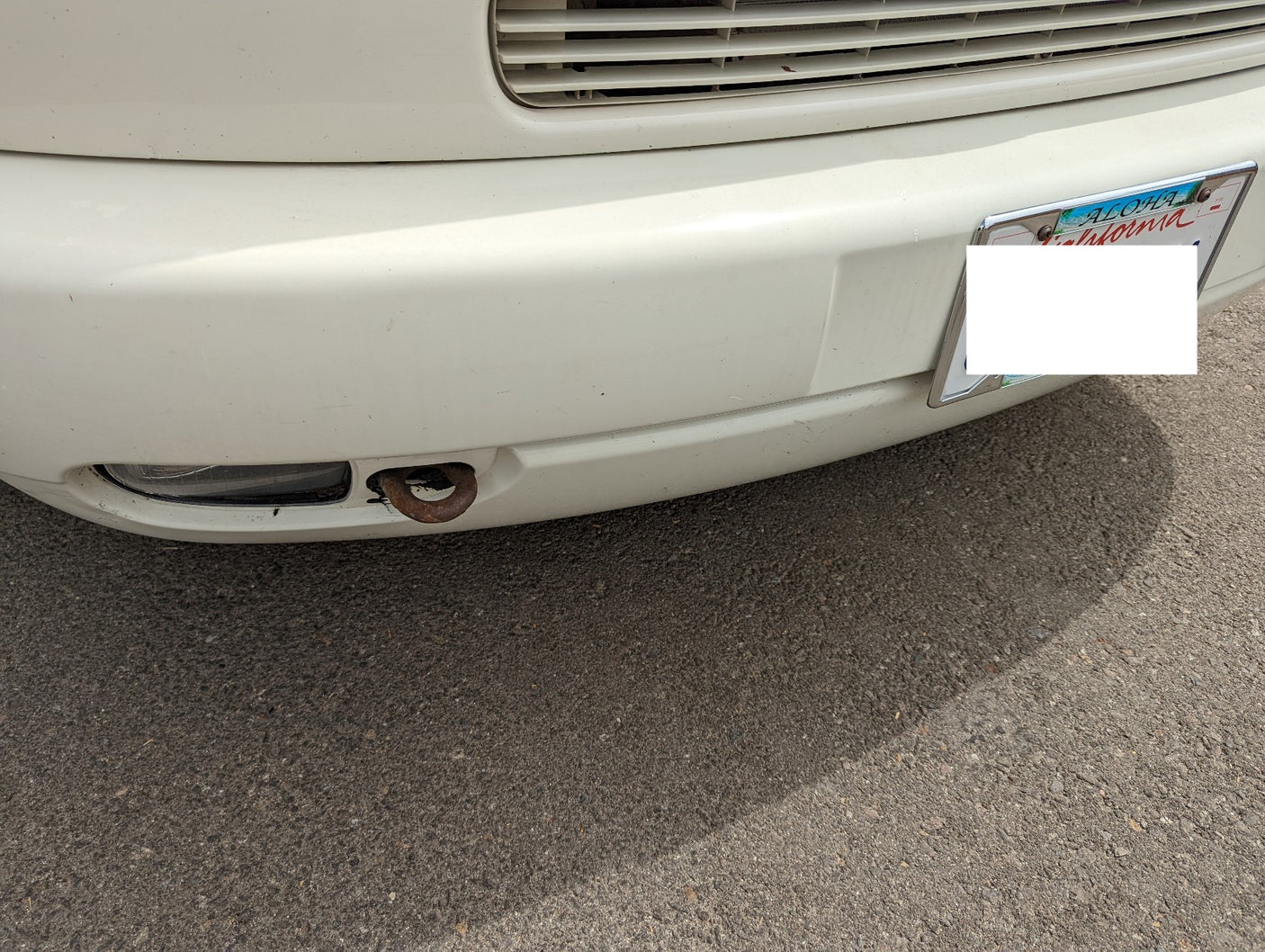 Front bumper of a white RV rental, showing a rusty tow hook and an 'Aloha California' license plate. Parked on asphalt.