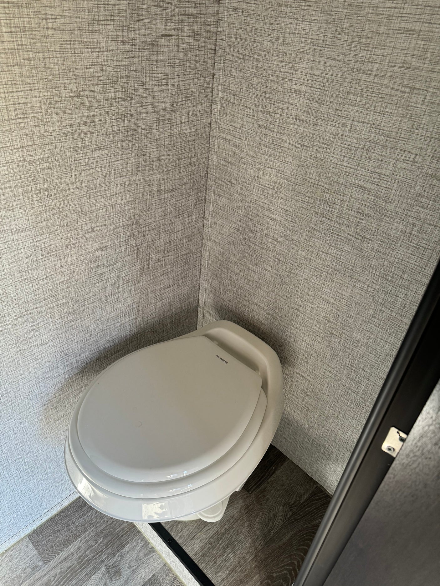 A white Dometic toilet in the compact bathroom of an RV rental, featuring textured gray walls and a grey wood-look floor.