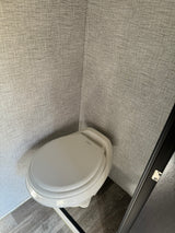A white Dometic toilet in the compact bathroom of an RV rental, featuring textured gray walls and a grey wood-look floor.