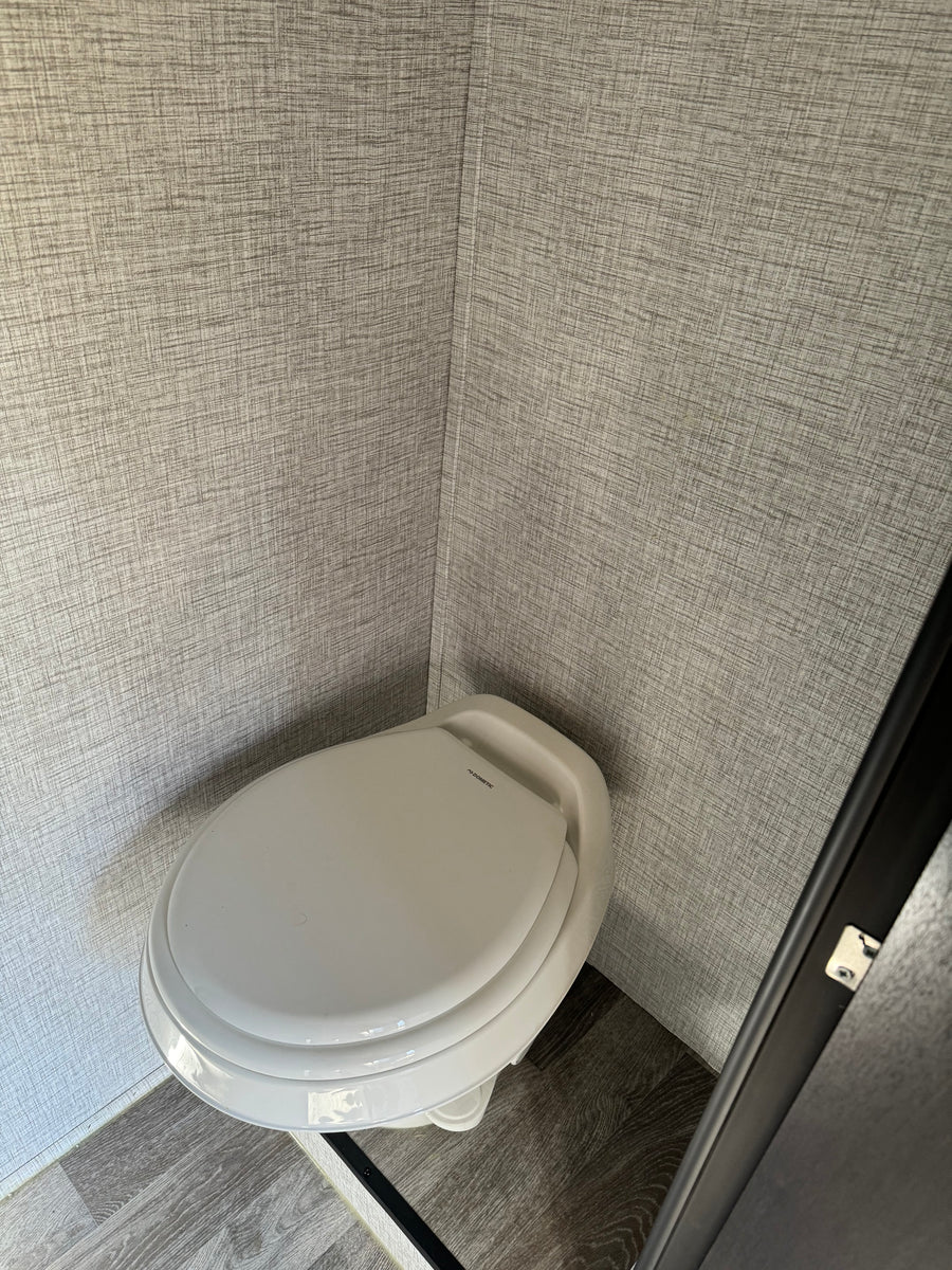 A white Dometic toilet in the compact bathroom of an RV rental, featuring textured gray walls and a grey wood-look floor.