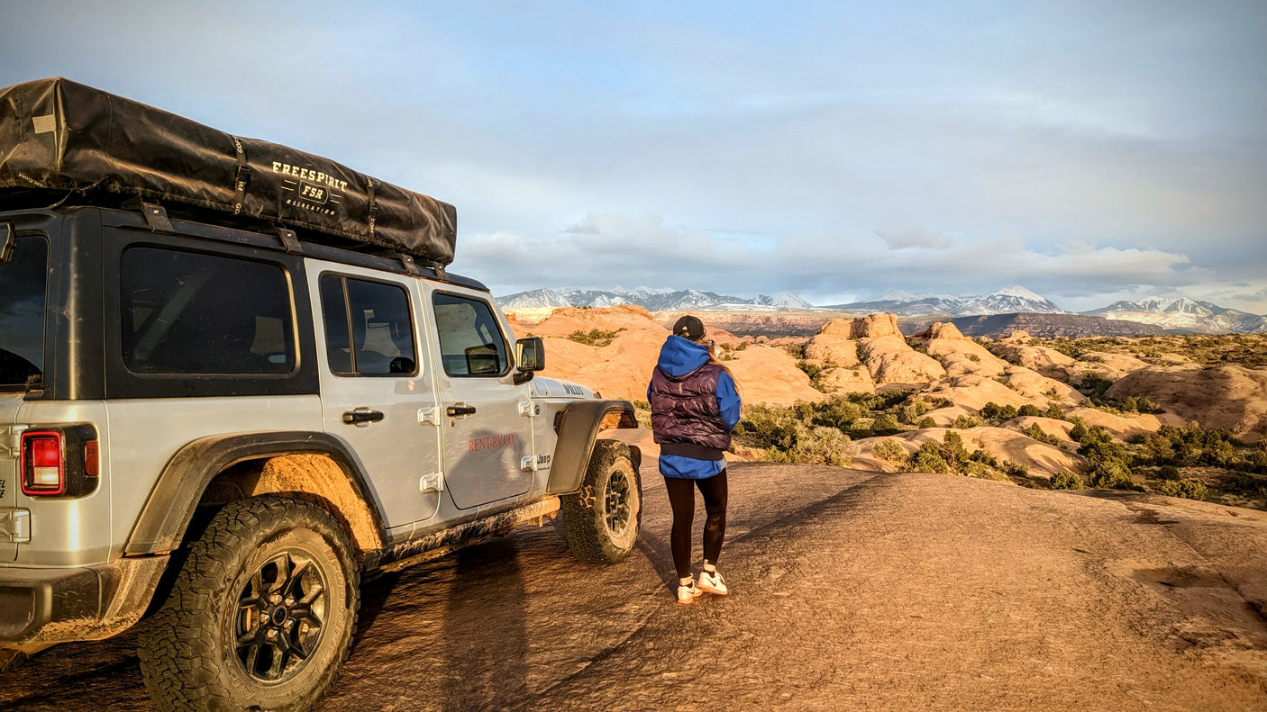 RV rental Jeep with rooftop tent on desert terrain. A person admires snow-capped mountain views.