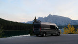 An RV rental camper van is parked by a calm lake with reflective trees and vast mountains under a pale sky. Perfect for nature escapes.