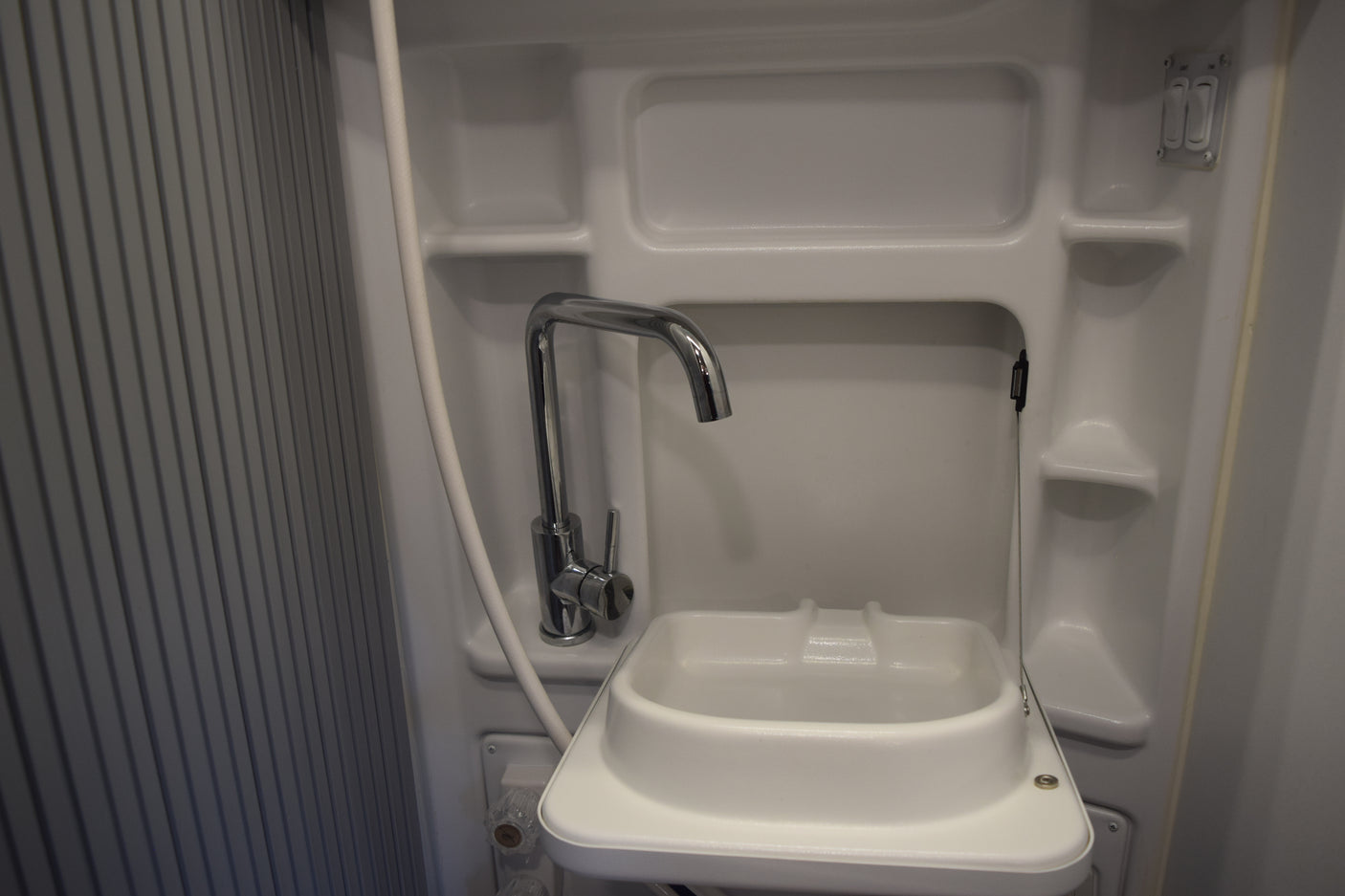 A compact, white RV rental bathroom featuring a sink, chrome faucet, attached shower hose, and built-in shelving.