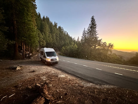 White RV rental van pulled over on a mountain road, surrounded by tall pine trees and a colorful sunset sky.