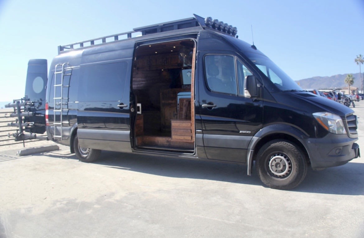  parked on a city street with buildings.|Black Mercedes Sprinter RV rental with roof rack and ladder — photo 1