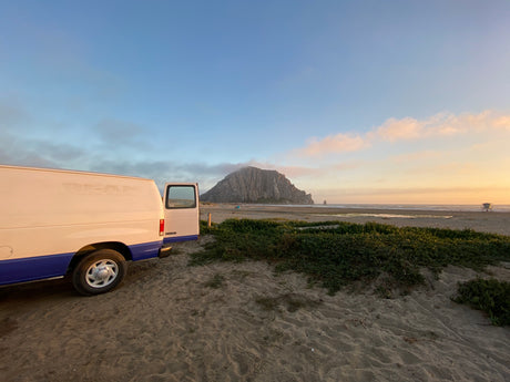 An RV rental van parked on a sandy beach, overlooking Morro Rock & the ocean at sunset.