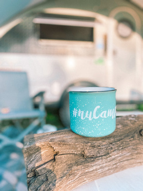 Cozy RV rental adventure: A turquoise #nuCamp mug sits on weathered wood, with a camper blurred behind. Ideal for camping.