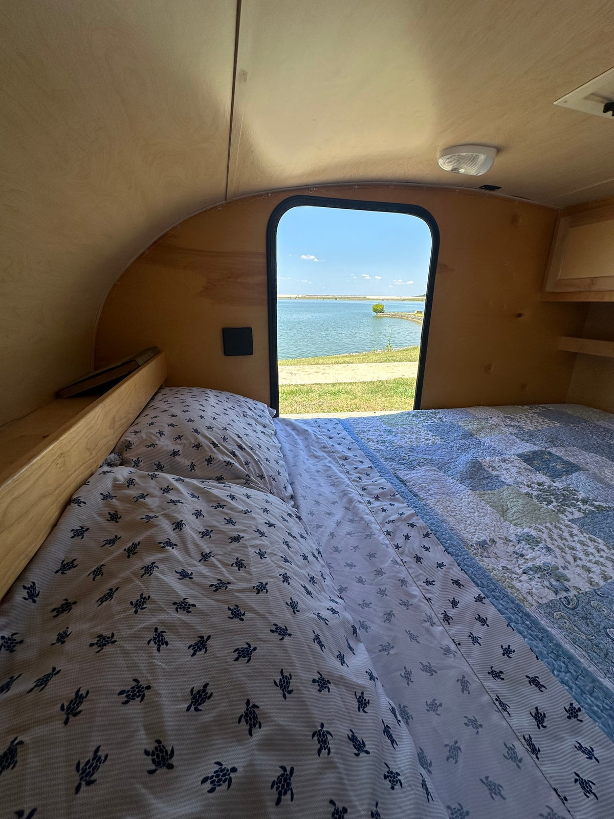  set against light wood paneling.|Open door of an RV rental camper showing interior storage pockets and turtle-patterned bedding.|Inside an RV rental teardrop camper: open door view of cozy bed with blue sea turtle sheets. Ready for adventure!|Cozy RV rental bed with turtle sheets and ambient string lights for a comfortable — photo 9
