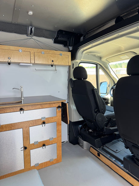 Custom RV rental interior showing a compact kitchenette with wooden cabinets and front seats.