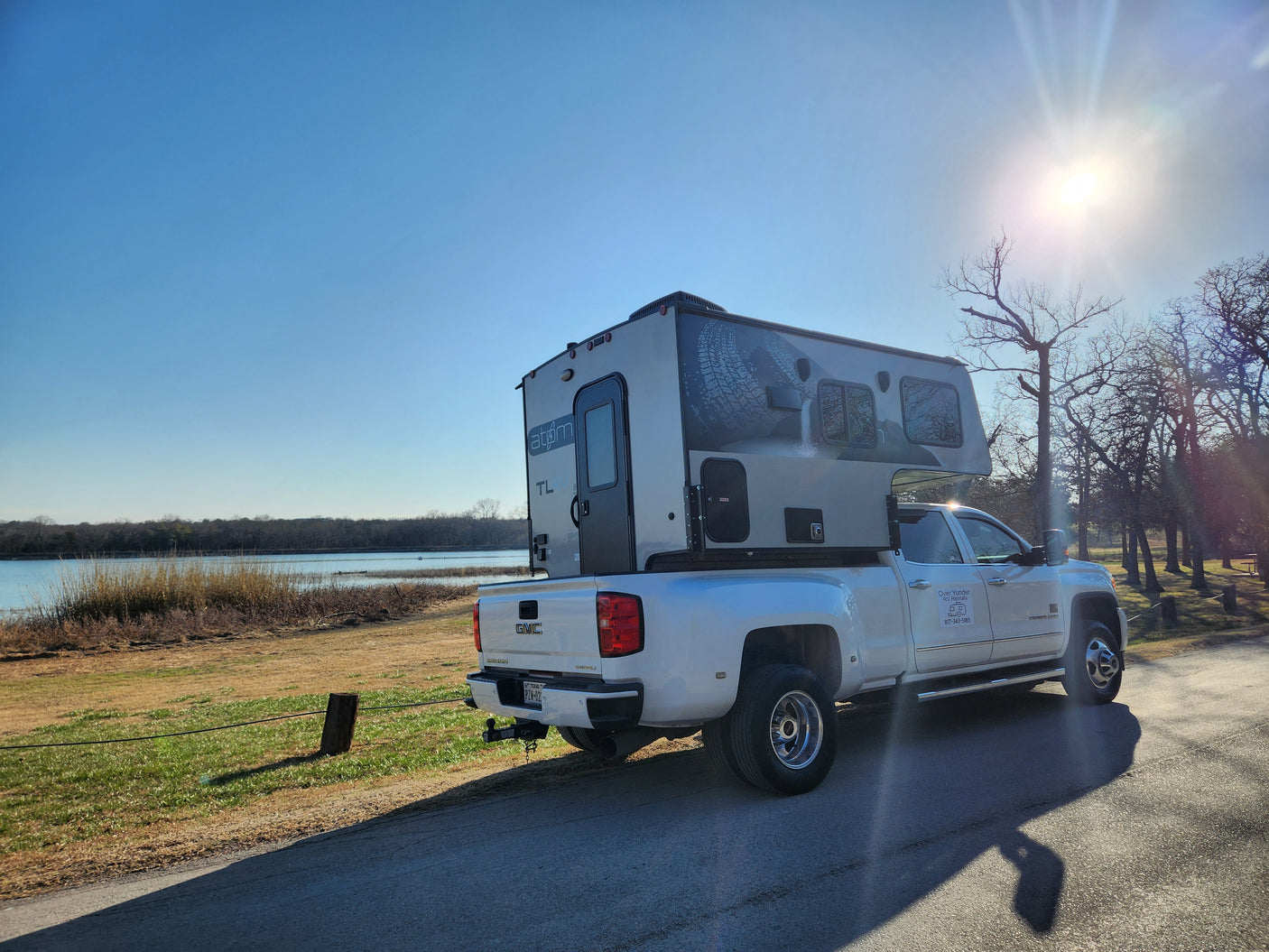White GMC dually truck with an Atom TL truck camper (RV rental) from Over Yonder, parked lakeside on a sunny day.