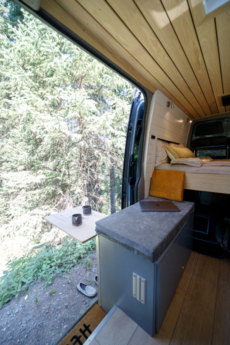 Enjoy nature from this cozy RV rental camper van with a wooden interior, bed, and pull-out table overlooking the forest.