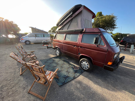 RV rental pop-top VW camper van & setup with chairs & rug at a sunny campsite.