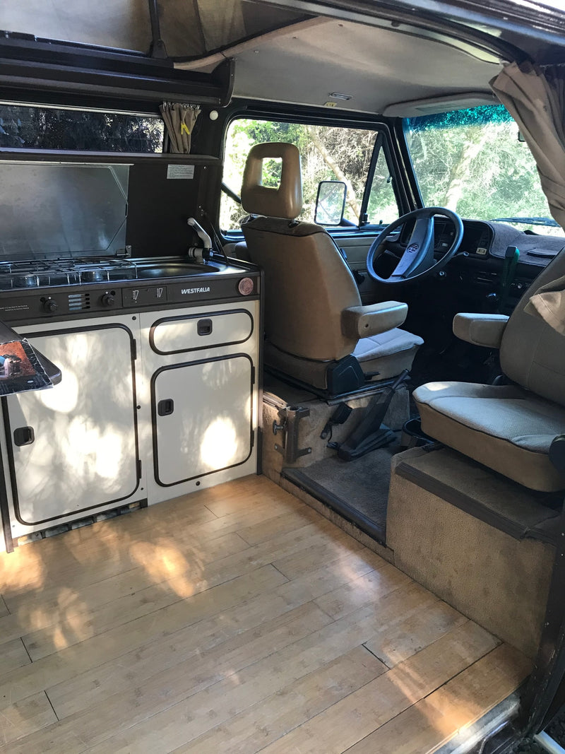  with fresh food & drinks ready on a table.|RV rental ready for adventure: Brown camper van with rear bike/cook station and outdoor dining table in a wooded camp.|Inside this RV rental: a Westfalia kitchen with stove & sink — photo 7