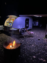 An illuminated teardrop rv rental camping setup at night, with an open kitchen, fire pit, and extended awning.