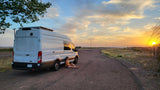 A person relaxes by their white RV rental camper van at sunset, a stunning golden glow over the quiet rural road.