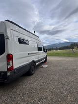 This RV rental camper van, parked on gravel, overlooks a field with horses and distant mountains under a cloudy sky.