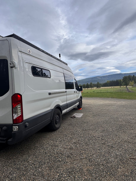 This RV rental camper van, parked on gravel, overlooks a field with horses and distant mountains under a cloudy sky.