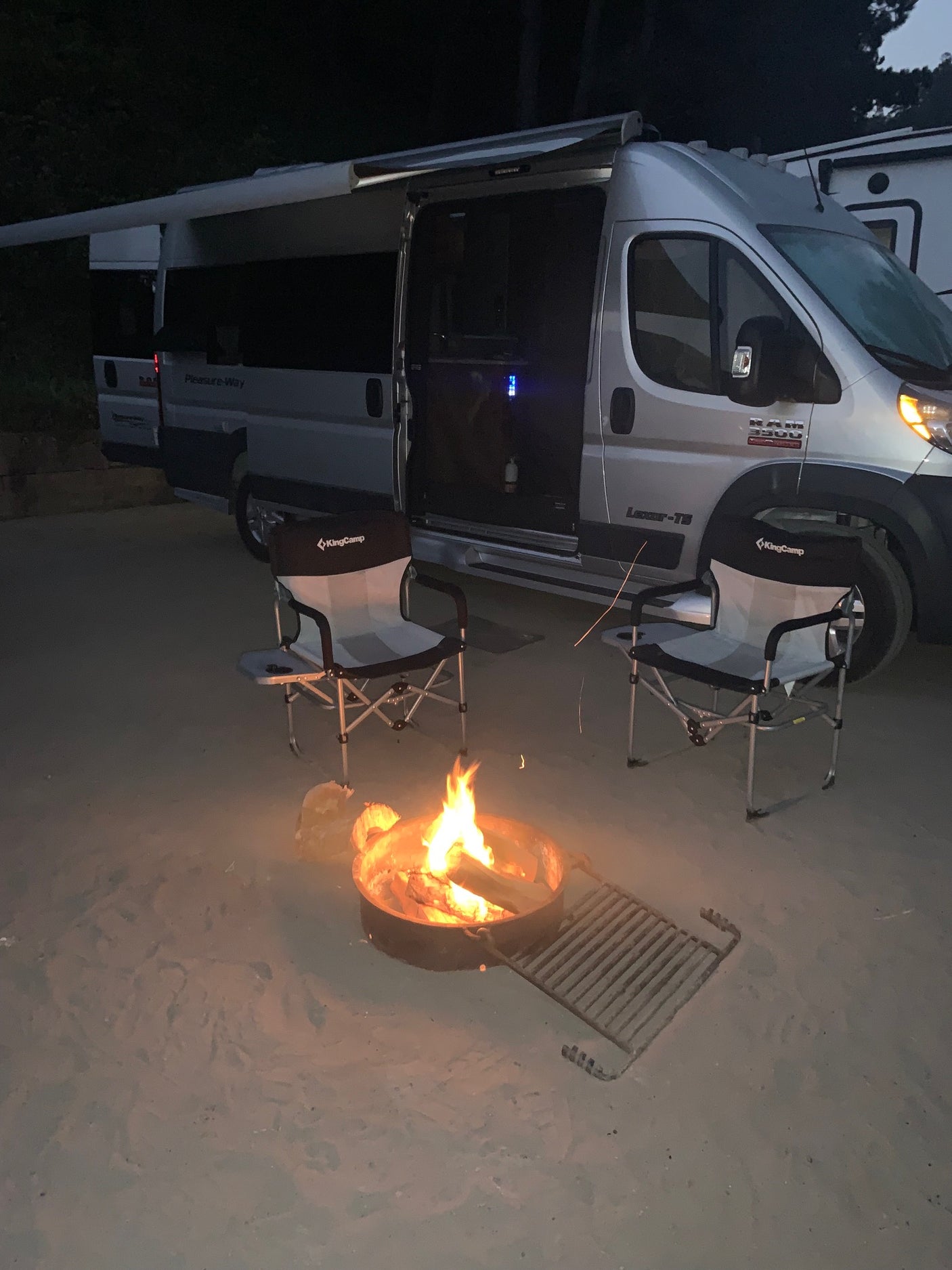 Cozy RV rental campsite at night: a silver campervan, two chairs, and a warm, glowing campfire on sandy ground.
