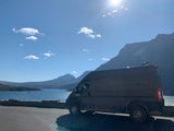 Dark RV rental van parked beside a stone wall, overlooking a vast blue lake & mountains under a bright sky.